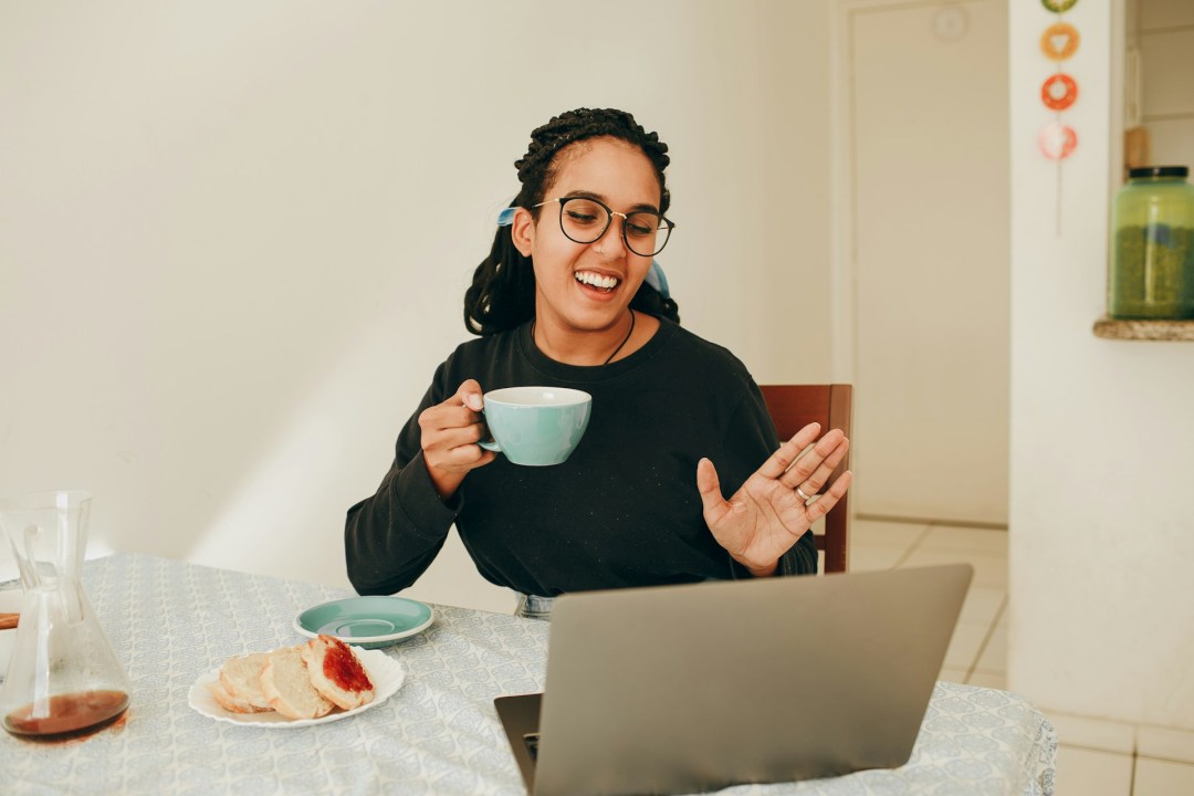 Woman with glasses holding a cup and waving during an online lesson on her laptop