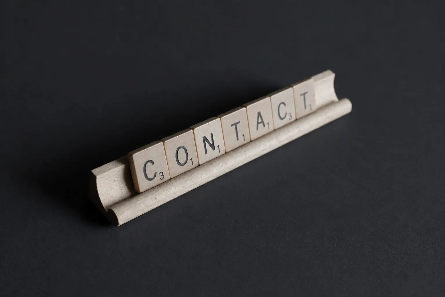 Scrabble tiles spelling contact on a wooden tile rack against an ink-grey background